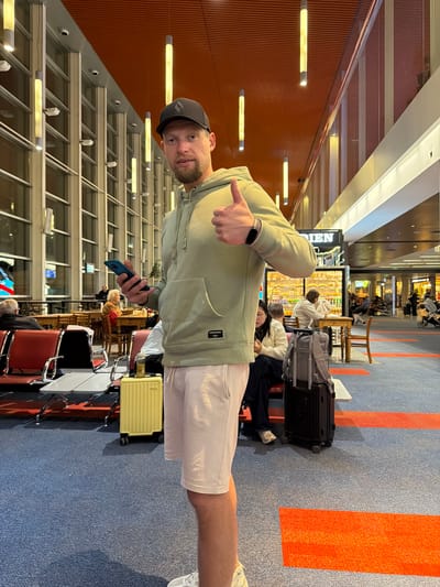 The image depicts a man standing in an airport terminal, possibly Aeropuerto Internacional Ezeiza in Argentina, based on the provided context. He is wearing a light green hoodie, light pink shorts, and a black baseball cap. The man is holding a blue smartphone in his left hand and giving a thumbs-up with his right hand. He is positioned in the foreground, with his body turned slightly towards the camera.

Behind the man, the airport environment is visible. There are rows of seating, luggage, and other travelers in the background. The terminal features a high ceiling with distinctive orange paneling and long, cylindrical light fixtures. Large windows line the left side of the image, revealing an outdoor area. The floor is carpeted in blue with red stripes. To the right of the man, there is a small yellow suitcase and a larger black suitcase with a gray backpack on top. In the middle ground, there are tables and chairs where other people are seated, some appearing to be eating or working on their phones. Further in the background, more seating areas and shops are visible. The overall atmosphere suggests a busy travel hub.