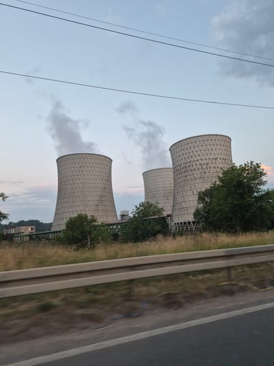 The image shows a power plant in Bukinje, Bosnia and Herzegovina, featuring three large cooling towers. Two of the cooling towers are actively emitting steam, with one of them the most prominent subject in the image. The towers are cylindrical and have a distinctive patterned exterior. In front of the cooling towers, there is a green pipe system supported by a metallic structure. The area surrounding the power plant is covered in dry grass and scattered trees. A road with a metal guardrail is visible in the foreground, suggesting the image was taken from a moving vehicle. The sky is overcast with some hints of pink and orange from what appears to be a sunset.