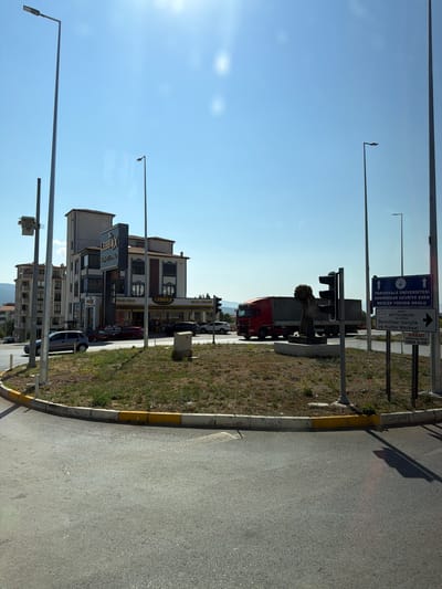 The image captures a sunny daytime scene at a traffic circle in Serinhisar, Turkey. In the background, a multi-story building with the "LEBBOX" signage is visible, along with other apartment buildings. A red semi-trailer truck is in motion, passing through the traffic circle, and several cars are parked or driving nearby. A traffic light stands next to a sculpture of a person holding what appears to be a bouquet of flowers. Road signs indicate directions to "PAMUKKALE UNIVERSITY SERINHISAR CEVRIYE ESEN MESLEK YUKSEK OKULU" and "DENIZLI BUYUKSEHIR BELEDIYESI SERINHISAR Gençlik ve Kültür Merkezi," as well as a warning sign for a "DIKKATI DİREKSİYON EĞİTİM SINAV ALANI" (Attention Driving Education Exam Area). Streetlights are present throughout the scene, and the ground is paved asphalt, with a grass-covered median in the center of the traffic circle, bordered by a yellow and white curb. The sky is clear blue.