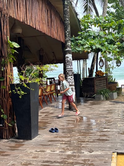 The image shows a child walking along a wooden boardwalk in Talpe, Sri Lanka, near a beach. The child is wearing a light-colored shirt, maroon shorts, and pink sandals. They are walking away from the camera, toward the beach. To the left of the boardwalk is a building with a wooden roof and walls. A decorated palm tree supports the roof. To the right are plants in containers. A pair of sunglasses rests on the boardwalk. The wooden planks of the boardwalk appear wet. In the background, the ocean is visible.