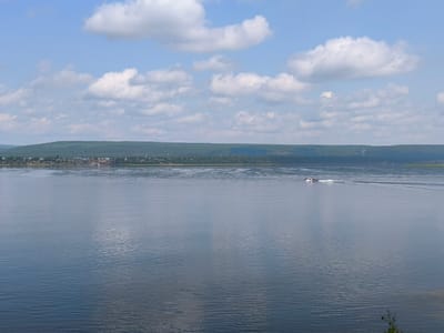 A wide, tranquil river flows through a landscape under a partially cloudy blue sky. The water surface reflects the sky and the distant shore, creating a mirror-like effect with subtle ripples. On the far bank, a dense forest lines the horizon, with a small settlement of buildings visible amidst the trees. A single motorboat is moving from left to right across the river, leaving a faint wake behind it. The overall scene suggests a peaceful day in a rural or semi-rural setting. The town of Boguchany is situated along the Angara River in Krasnoyarsk Krai, Russia, and this image likely depicts a view of the Angara River near the town.