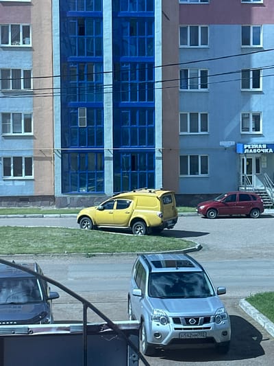 The image depicts a street scene in Mariinsky, Russia, featuring an apartment building with distinct architectural elements. The building's facade is a mix of light blue and beige, contrasted with vibrant blue windows. Several cars are visible: a yellow pickup truck with a camper shell is parked on a patch of grass, a red hatchback is parked on the asphalt, and a silver SUV is parked in the foreground. A sign above a storefront on the building reads "РЕЗНАЯ ЛАВОЧКА," indicating a shop. Power lines crisscross the scene in front of the building. The sky is partly cloudy, casting shadows on the cars and ground.