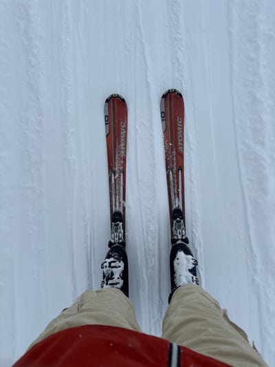 A first-person, downward-facing view captures a skier's lower extremities on a groomed slope in Bad Gastein, Austria. The individual is wearing tan ski pants, with a partial red garment visible at the bottom of the frame. Two black ski boots are securely fastened into bindings on a pair of red "ATOMIC" brand skis, each marked with "10". Accumulations of white snow are visible on both the boots and the skis. The snow-covered terrain features distinct parallel tracks, indicating recent grooming. The scene depicts a stationary individual equipped for skiing on a prepared winter sports surface.