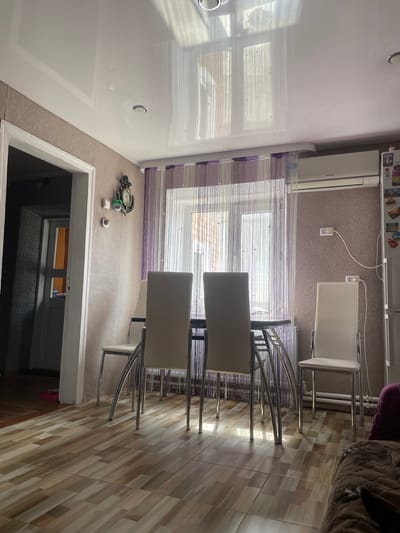 The image shows an interior scene of a room, likely a dining area or kitchen, with a dining table and chairs. The room has tiled flooring and a light-colored, possibly stretch fabric, ceiling with visible lighting fixtures. A window is covered with a beaded curtain. On the wall, there is a decorative clock with an anchor motif and what appears to be a Samsung air conditioning unit. There is also a refrigerator visible on the right side of the image. The overall impression is that of a lived-in residential space. There are no people visible in the image, nor any discernible actions or interactions taking place. The location is identified as being in the city of Salsk, Russia.