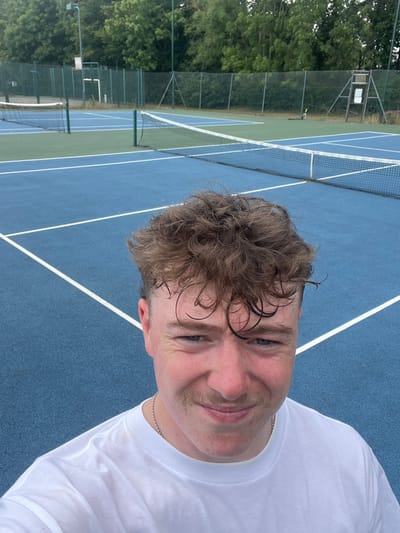 A young adult male is depicted in a close-up, frontal view, occupying the lower portion of the image. He has light brown, wet-looking curly hair, a light mustache, and blue eyes. He wears a white T-shirt and a silver ball-chain necklace, looking forward with a slight upward turn to the lips. Behind him are two outdoor tennis courts. The immediate background shows a blue tennis court with white lines and a green net. Beyond this, a second, parallel court is visible, also blue with white lines and a green net, transitioning to a green perimeter. A green chain-link fence surrounds the courts, backed by dense green foliage. A tall light standard is on the left, and a sign structure is on the right, both outside the fence. A tennis racket lies on the far court surface. The scene is located in Cherry Burton, United Kingdom.