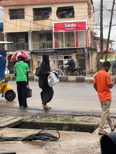 In an outdoor urban setting, under a cloudy sky, a group of people are walking and riding on a street. A yellow motorcycle is parked on the left side of the road. A woman in a green shirt and black pants walks away from the camera, carrying a bag in her right hand. Next to her, a woman wearing a black hijab and abaya walks towards the camera, carrying a large, crinkled silver bag in her right hand. Further down the street, a man rides a motorcycle with a passenger behind him. On the right side of the frame, a young man in an orange t-shirt and khaki pants walks away from the camera, looking at his phone. In the background, a multi-story building with shops on the ground floor is visible. Power lines crisscross the sky above the street. The street is wet, suggesting it has recently rained, and a drainage ditch runs along the side of the road, with black cables coiled within it.