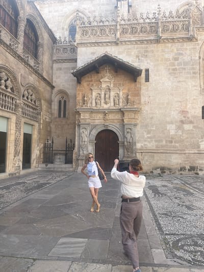 An eye-level, full outdoor shot shows two women standing in a stone-paved plaza in front of a large, ornate building in Granada, Spain. The building features intricate stone carvings around its arched doorway and windows. One woman, on the left, faces the camera, posing with her hands on her hips. She wears a striped blue and white shirt, white shorts, gold sandals, sunglasses, and a white scarf around her neck. The second woman, on the right, stands with her back to the camera, holding up a phone to take a photo of the first woman. She is wearing a white long-sleeved shirt, brown and white pinstriped pants, and a red scarf around her neck. The plaza floor is a mosaic of different stone patterns, with one section featuring a detailed animal mosaic on the right side of the frame. The building appears to be an old church or cathedral, characteristic of the architecture found in Granada.
