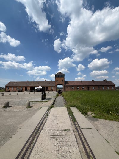 This media file is a photograph taken outdoors on a sunny day with scattered white clouds against a vibrant blue sky. The image is oriented vertically, suggesting it might have been captured on a phone.
The main subject is a long, multi-story brick building with a red tile roof and numerous evenly spaced windows. To the right of the building, there is a patch of neatly trimmed green grass. In front of the building, there is a paved area made of concrete and gravel, with what appear to be railway tracks embedded in the concrete. The tracks run alongside the paved area, disappearing into the distance.
In the foreground, on the paved area, there are several round, grey concrete bollards. A portion of another brick structure with a tall tower is visible on the left side of the frame, extending over an arched entrance. A small group of people, dressed in casual clothing, are visible near this entrance and further down the paved area. Some of them appear to be looking around or walking. One person is standing near a small white umbrella structure, possibly a refreshment stand or information point.
The overall scene suggests a historical site or memorial, possibly a former camp or industrial facility, given the presence of barracks-like buildings and railway tracks. The bright sunlight and clear sky indicate it is daytime, likely late morning or afternoon. The neat grass and well-maintained appearance of the buildings suggest it is an active site, perhaps a museum or memorial park. There is no visible text in the image. The location context provided is Brzezinka, Poland, which is historically significant as the site of the Auschwitz-Birkenau concentration and extermination camp.