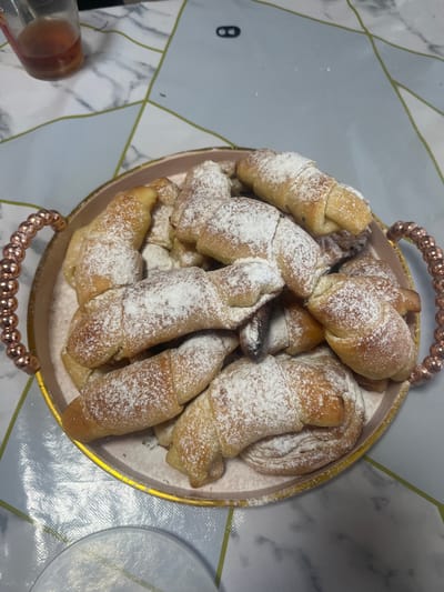 In Oktyabrskoye, Kazakhstan, a pink-rimmed tray filled with crescent-shaped pastries dusted with powdered sugar sits on a patterned tablecloth. A glass with a reddish liquid is visible in the upper left corner.