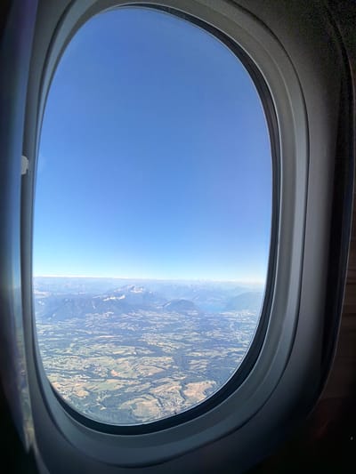 This media file shows a view from an airplane window looking out at the sky and a landscape below. The window frame is dark gray and oval-shaped, with a lighter gray inner rim. The sky is a clear, bright blue, filling the left side of the frame. On the right side, a landscape is visible, appearing to be a rural area with rolling hills, patchwork fields in various shades of green and brown, and scattered clusters of buildings. The landscape suggests a geographic location with varied terrain, possibly mountainous due to the texture of the land.
The scene suggests the airplane is in flight during the daytime, with the bright blue sky and clear visibility indicating fair weather. The horizon line where the sky meets the land is sharp and defined, further supporting good weather conditions. The perspective from above implies the photo was taken from a significant altitude.
While no people are directly visible, the context of an airplane window implies the presence of passengers. The activity taking place is travel by air. The overall mood conveyed is one of tranquility and the wonder of flight, with the vast expanse of the sky and the detailed view of the earth below.
There is no visible text in the media file. The mention of "Bourg-Saint-Maurice, France" provides a geographical context for the landscape depicted.