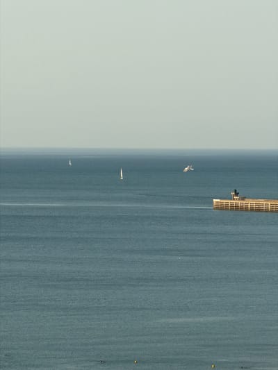 This aerial, vertical image depicts a wide expanse of calm, blue water, likely the sea, with a thin, hazy layer of atmosphere at the top left. A pier or jetty stretches horizontally across the upper portion of the frame, characterized by a grid-like structure and an attached, dark, box-like element.
Several small sailboats are visible on the water. One is closer to the pier, appearing as a white triangular sail. Further down, another sailboat is visible, also with a white sail. A third, smaller sailboat is even further away. A single seagull is captured in flight between the pier and the sailboats, its wings spread. Small, yellow buoys are dotted along the right edge of the water.
The lighting suggests it is daytime, possibly late afternoon or early morning given the soft, diffused light and the muted colors. The water appears still, indicating calm weather conditions. The presence of the pier and the sailboats suggests a coastal location where recreational sailing is common, consistent with the provided location context of Dieppe, France. No text is visible in the image.