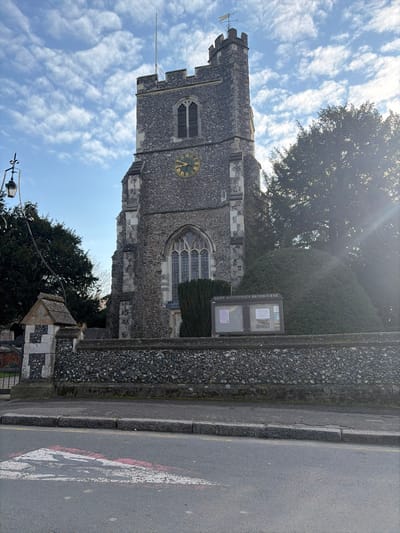 The image depicts St. Augustine's Church in Broxbourne, United Kingdom, viewed from across a street. The prominent feature is the church's stone tower, constructed from grey, irregular stone blocks. A rectangular clock face with gold numbers and hands on a green background is visible on the tower, positioned below a louvered belfry window. The tower is topped with battlements, a flagpole, and a weathervane. Below the clock, a large, arched, multi-paned Gothic-style window is present.
In the foreground, a low, rugged stone wall, matching the church's material, separates the churchyard from the street. To the left, an elaborate stone gatepost with a small, gabled roof structure and a black ornamental street lamp marks an entrance. An information board displaying "SAINT AUGUSTINE'S BROXBOURNE" is situated behind the wall to the right of the gatepost. Dense green foliage, including tall trees and a shaped bush, partially frames the church.
The immediate foreground shows a grey asphalt road with a curb and pavement, and faded red and white markings are visible on the road surface. The sky is bright blue with scattered white clouds, and sunlight illuminates the scene, creating a lens flare on the right side of the image. No individuals or vehicles are present.
