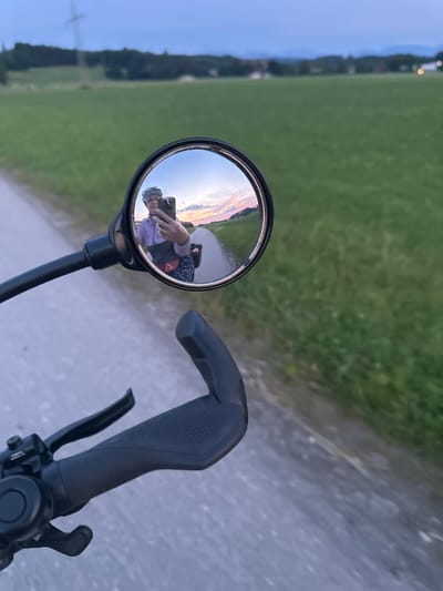 The image is a first-person perspective of someone riding a bicycle, captured by a camera attached to the handlebars and angled to include a rearview mirror. The scene shows a paved path running through a grassy field. In the distance, trees line the horizon, and the sky suggests it is either dawn or dusk, with soft, warm colors reflecting in the mirror. The rearview mirror itself shows a reflection of the rider, who is wearing a helmet and a light purple jacket. The rider is holding a smartphone in their hand, likely to take the photo or record a video. The handlebars, with their grip and brake levers, are prominently in the foreground. The lighting and the colors in the sky indicate a peaceful, possibly serene moment during a bicycle ride in the German countryside, near Bad Endorf. There is no visible text in the image.