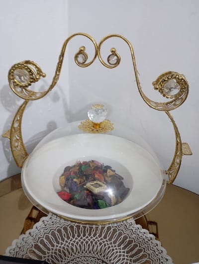 The image displays a decorative centerpiece, likely a potpourri holder or a covered serving dish, set against a plain, light-colored wall. The central object features a white ceramic or porcelain bowl, filled with colorful potpourri, and covered by a clear, transparent dome lid.
The bowl rests within an elaborate, golden-colored metal stand. This stand is highly ornate, characterized by intricate scrollwork and decorative cut-out patterns along its curved supports. Two main supports arch gracefully from a base (partially obscured by a doily) over the central bowl, meeting at a point near the top. Each support is adorned with clear, faceted crystal-like embellishments: smaller, round crystals are set into the upper, inward-facing curls of the stand, while larger, oval-shaped crystals are prominently featured within more intricate, swirling metalwork at the outer, broader curves of the supports. A small, golden, intricately designed finial with a faceted crystal knob secures the clear dome lid at its apex.
Inside the clear-domed bowl, a collection of dried botanicals, commonly known as potpourri, is visible. These pieces vary in color, including shades of dark green, reddish-brown, purple, orange, and lighter beige or tan, suggesting a mix of dried leaves, petals, and seed pods. Among the potpourri, a small, golden, rectangular decorative item with a textured or framed appearance can be seen.
The entire decorative piece is placed on a highly reflective, shiny golden surface, possibly a tray or a polished table. Beneath the stand and extending towards the viewer is a light brown or beige textile doily with an elaborate, circular, open-work pattern, resembling lace or crochet. The background is a simple, off-white or light gray wall, with a subtle corner or seam visible on the left side, indicating an indoor setting. The lighting is even and bright, highlighting the metallic sheen and crystal details of the centerpiece. There are no people, visible activities, or discernible text in the image.