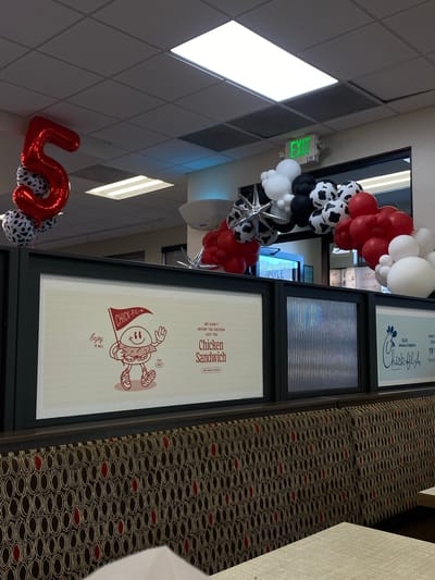 The image shows a brightly lit interior of a Chick-fil-A restaurant in Hagerstown, United States of America, likely decorated for a celebration. In the foreground, a patterned booth with brown, black, and red circular designs lines the seating area. Above the booth, a large banner displays a cartoon chicken character holding a flag that says "CHICK-FIL-A" with the slogan "Enjoy IT ALL". To the right of this banner, another smaller banner features the Chick-fil-A logo and text.

Dominating the background is a balloon arch in red, white, and black with cow-print patterned balloons, and a large, red "5" balloon. An "EXIT" sign glows green above the balloon display. The ceiling is a typical drop ceiling with fluorescent lighting. The overall scene suggests a festive atmosphere within the restaurant.