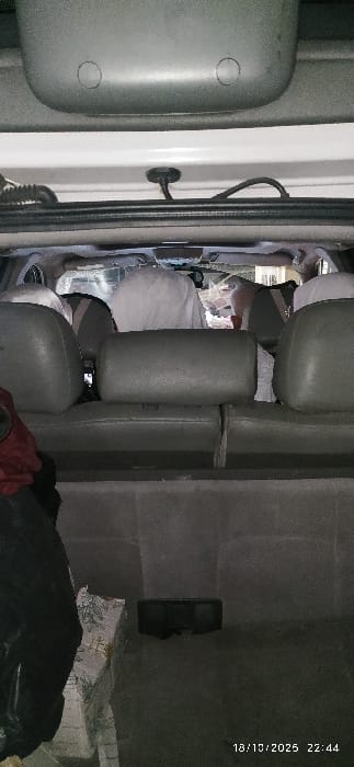 The scene is set inside a vehicle located in the city of Al-Kiswa, Syria, viewed from the rear cargo area looking forward into the passenger compartment. The image was captured at 22:44 on October 18, 2025, in low light conditions. The vehicle's interior features a white ceiling and hatch frame, with a gray plastic overhead component containing two circular indentations. Exposed black wiring and a corrugated conduit are visible near the rear roofline.

In the foreground, a dark gray upholstered bench seat with three headrests faces forward. Beyond this seat, multiple individuals are seated in subsequent rows. At least three figures are discernible, largely obscured by white fabric or garments covering their heads and upper bodies. One figure is centrally positioned behind the foreground seat's middle headrest. Distant, indistinct lights or reflections are visible through the vehicle's front windshield, indicating an outdoor setting.

The cargo area floor is dark gray. On the left, a large item is wrapped in dark material, with a portion of a lighter, patterned object visible beneath or next to it. A dark, rectangular object, possibly an electronic device, is positioned on the floor towards the center-right.