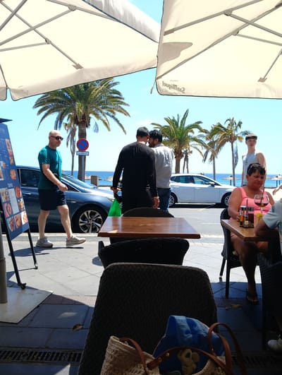 This media file captures a vibrant, sunny outdoor scene at a coastal cafe in what appears to be Puerto Del Carmen, Spain. The image is taken from a low perspective, looking out from under the shade of two large white umbrellas, revealing a bustling street, palm trees, and the clear blue ocean with a beach in the background.
In the foreground, a person is seated at a wooden cafe table, wearing a pink top and holding a glass with a dark red beverage, possibly a cocktail or red wine. Sunglasses are perched on her head, and her expression is neutral. To her left, part of another person and table are visible. In the immediate foreground, a woven bag contains a blue cloth and a light-colored toy, possibly an elephant.
Across the sidewalk, several individuals are visible. A bald man wearing a green t-shirt, dark shorts, and sunglasses walks casually from right to left. Further along the street, two men are standing, one with his back to the camera wearing a black long-sleeved shirt, and another in a light grey shirt. A young man in a white tank top and a flat cap stands further down, looking towards the sea.
The street features a dark blue sedan and a white hatchback. Palm trees line the roadside, and a 'No Parking' sign, clearly displaying its universal symbol and some Spanish text like "CARGA Y DESCARGA" (loading and unloading), stands next to a tree. The background showcases the expansive blue sea and a sandy beach with what appear to be sun loungers or umbrellas. The cafe's menu board, featuring "Tapas" and other dishes, is prominent on the left. The umbrellas overhead bear the mirrored text "San Miguel CERVEZA," indicating a brand of beer. The bright sunlight and clear shadows suggest it's midday or early afternoon, with the weather being very clear and warm.