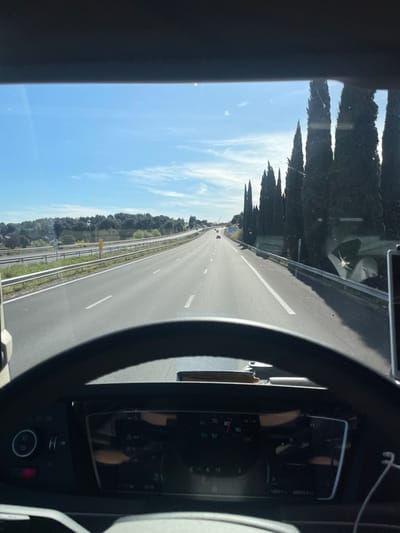 The image presents a forward-facing view from inside a vehicle, likely a truck, traversing a multi-lane highway under a clear blue sky with scattered cirrus clouds. The location is near Tavel, France.
In the immediate foreground, the top of a black steering wheel is visible, centered at the bottom of the frame. Below the steering wheel, a dark digital dashboard instrument cluster displays various readouts, including an odometer showing approximately 193,311 kilometers, speed or RPM indications (e.g., "70 80 90 100"), and other symbols such as "E," "A," and "12." A circular control knob and a red indicator light are visible to the left of the dashboard display. To the right, a white earbud with its cable hangs. A light brown object, possibly a wallet or document, rests on the dashboard surface below the windshield.
The highway, appearing dry and paved with asphalt, extends directly into the distance, marked by white dashed and solid lines. Multiple lanes are visible, accommodating vehicles traveling in the same direction; several indistinct vehicles are observable at a distance. A metal guardrail runs along the immediate right edge of the highway. Behind this guardrail, a dense row of tall, slender, dark green cypress trees lines the road, extending towards the horizon and casting shadows. On the left side of the highway, another metal guardrail separates the main roadway from an elevated bank covered in lush green vegetation and trees. A yellow road sign or marker is partially visible on the left side. The scene is illuminated by bright daylight, indicating clear weather conditions.