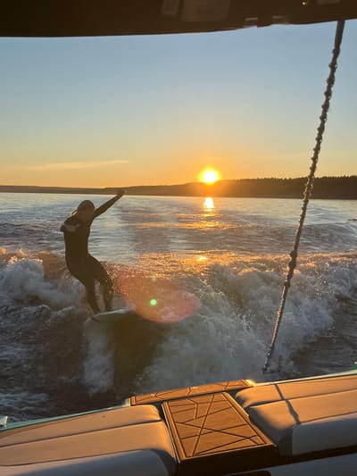 A person in a black wetsuit surfs a wave behind a boat at sunset. The sun is low on the horizon, casting a warm glow across the water. The boat's wake creates a large, foamy wave for the surfer. The scene appears to be taking place on a lake or calm body of water, with a tree-lined shore visible in the distance. The image is taken from the perspective of someone on the boat, looking back at the surfer. The location is Gromovo, Russia.
