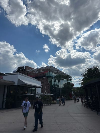 The image shows an outdoor scene in Lake Buena Vista, Florida, United States, likely near Disney Springs. The sky is bright blue with fluffy white clouds. A building with a Coca-Cola store and beverage bar sign is partially visible. People are walking along a paved pathway. Two men are in the foreground, one wearing a white t-shirt and shorts, and the other wearing a dark t-shirt and jeans. There are trees and the edge of a covered structure on the right side of the frame. Other people walk on the paved path further back, and there are tables and chairs under the structure. The building has a brick facade and glass elements.