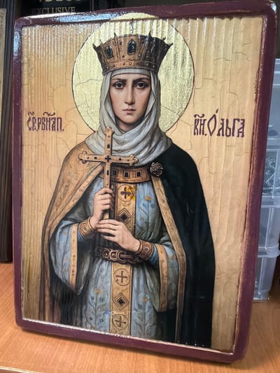 This media file depicts an icon painted on wood, featuring Saint Olga. She is shown in a traditional Byzantine style, with a halo and a crown, signifying her royal status. She is dressed in royal regalia, including a blue robe adorned with intricate patterns and a dark cloak. In her hands, she holds a cross, a symbol of her Christian faith and her role as a saint.
The icon is set against a gold background, typical of religious iconography, which symbolizes divine light. The wood panel shows signs of age and cracking, adding to the antique and revered nature of the object. Visible text in Cyrillic script appears in two locations on the icon. At the top, above Saint Olga's head, the text reads "КН. ОЛЬГА" (KN. OLGA), likely referring to her title as Princess Olga. Below her, on the lower part of the icon, the text reads "СЕРПУХО" (SERPUKHO), which might be a signature or a place of origin, potentially referencing Serpukhov, a city historically significant in Russia.
The overall mood conveyed by the icon is solemn and devotional. The saint's expression is serious and serene. The setting is implied to be religious or historical, given the subject matter and style. No specific time of day or weather conditions are depicted as this is a religious artwork rather than a scene from everyday life. The context provided indicates the location as Maloyaroslavets, Russia, which is consistent with the Russian text and religious subject matter.