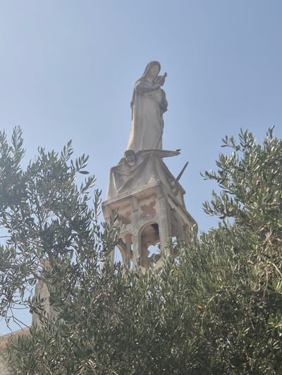 The image shows a statue of Mary and the infant Jesus atop a stone structure, partially obscured by olive tree branches. The statue is made of a light-colored material, possibly stone or concrete, and depicts Mary with a veiled head, holding a baby. The structure below the statue appears to be a bell tower or a similar architectural element, with arched openings and decorative stonework. A small bird is perched on the edge of the structure near the statue. The background is a clear blue sky, suggesting an outdoor setting. The presence of olive trees indicates a Mediterranean climate. This statue is located in the city of Abu Gosh, Israel, a town known for its Arab Christian community and religious sites.