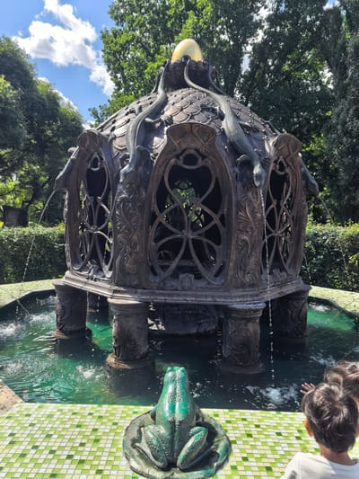 In this media file, a boy and another child are looking at a large, ornate fountain in a garden. The fountain, made of dark metal, has a domed roof covered in scales and is adorned with dragon-like figures. Water spouts from the mouths of these figures, cascading into a pool of green water. The fountain sits on four decorative columns. In the foreground, a green frog sculpture sits on a lily pad in a shallow section of the pool, which is lined with light green and white tiles. Lush green trees and bushes surround the fountain, indicating an outdoor setting with ample vegetation. The sky above is blue with scattered white clouds, suggesting it is daytime and the weather is pleasant. The presence of children suggests a recreational or tourist location. The image appears to be taken in the Sovkhoza imeni Lenina, Russia, as per the provided location context.