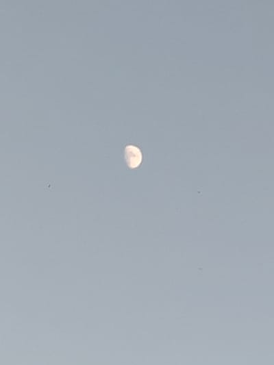 The image shows a pale blue sky with a small, crescent-shaped object in the upper center. The object is lit from the left and appears to be the moon or a similar celestial body. There are two very small dark spots visible in the sky, one to the left and slightly below the crescent object, and another to the right and slightly below the crescent object. The location is identified as Воўчынскі сельскі Савет, Belarus.