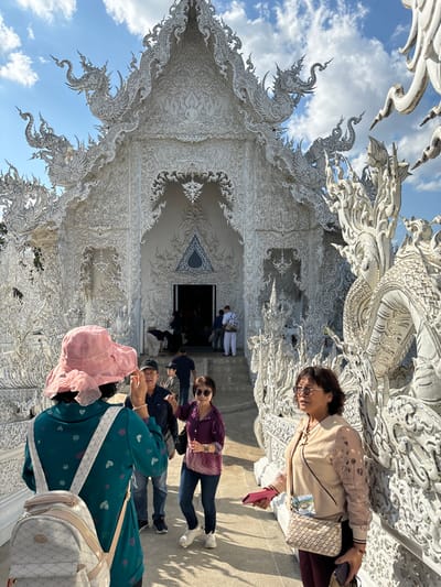 The image displays the Wat Rong Khun (White Temple) in Ban Rong Khun, Thailand, under a partly cloudy blue sky. The temple's main hall, rendered entirely in white with elaborate, multi-tiered architecture and intricate sculptural details including mythical creatures, occupies the background. In the immediate foreground, a person faces away from the camera, wearing a pink wide-brimmed hat, a teal long-sleeved shirt, and a white backpack. Their left hand is raised. To their right, a man in a dark baseball cap and shirt smiles and looks towards the right. Adjacent to him, a woman in a purple and pink long-sleeved top, jeans, and white sneakers, with sunglasses resting on her head, also smiles and looks right while holding her left arm bent. Further to the right, a woman with sunglasses on her face, dressed in a beige long-sleeved top, dark pants, and carrying a patterned crossbody bag, holds a pink object and a dark purse as she looks towards the right. Several less distinct individuals are visible ascending the steps into the dark entrance of the temple in the mid-background. The scene is brightly illuminated by sunlight, casting shadows on the light-colored ground.