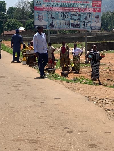 The image depicts a street scene in Kukum, Nigeria. A large political billboard stands beside the road featuring pictures of people and Hausa script. Several people stand beside the road selling various wares, including fruit. The road is paved and the surrounding environment is dirt and vegetation.