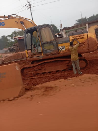 An excavator is positioned on a dirt road under an overcast sky. A man in a tan shirt and grey pants is seen facing away from the camera, interacting with the side of the excavator. The excavator is yellow and black with "5-EX12 XE2150" visible on its side. The ground is covered in reddish-brown dirt, suggesting a construction or excavation site. The background shows a few buildings and trees. The image appears to be taken in Imilike, Nigeria.