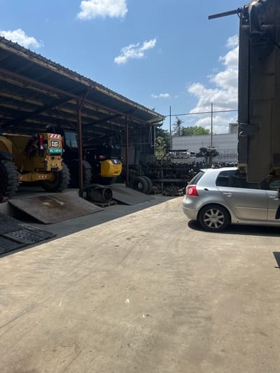 The image shows an outdoor junkyard or salvage yard under a clear blue sky with scattered clouds, likely in Hotantsa, Bulgaria. The scene is dominated by various vehicles and machinery, suggesting a place where old or damaged vehicles are stored and disassembled for parts.
In the foreground, a yellow Caterpillar (CAT) construction vehicle, possibly a loader, is parked. It has large tires and the CAT logo is clearly visible on its side. Next to it, a smaller yellow piece of heavy machinery is also visible. Behind these, a dark-colored vehicle is partially obscured.
To the right of the construction equipment, a silver Volkswagen car is parked on a concrete surface. It appears to be a hatchback model. Further into the background, shelves filled with automotive parts and more machinery are stacked. The overall setting is industrial and utilitarian, with a metal roof structure providing some shade over parts of the yard.
The lighting suggests it is daytime, likely late morning or afternoon, given the bright sunlight and distinct shadows. The weather appears to be fair and warm. There is no visible activity or people in the frame, implying the junkyard is either closed or not currently in operation. The image conveys a sense of disarray and obsolescence, typical of a vehicle salvage yard. The license plate on the CAT vehicle reads "WD14MPVC".