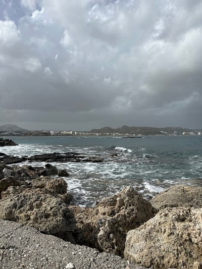 The image depicts a coastline near Faliraki, Greece, on a cloudy day. Large, rough, light-colored rocks form the immediate foreground, transitioning into a darker, more jagged rocky shoreline where the sea meets the land. Waves crash against the rocks, creating white foam.
In the midground is the sea, a gray-blue color, stretching towards a distant landmass. A town or city sits along the coastline of the distant land, with buildings of varying heights visible.
The sky is heavily overcast with dark, gray clouds filling the majority of the upper portion of the image. The lighting is dim and diffused.