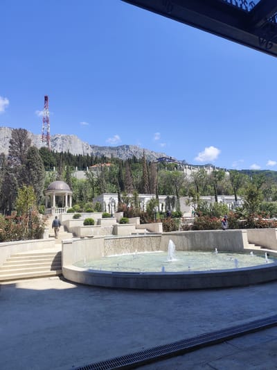 The image shows a tiered fountain in a landscaped garden. The fountain is circular with multiple jets of water spraying upwards. Water also cascades over the edges of the upper tiers into the fountain below. A set of steps leads up to a gazebo located on a higher level, with a person walking towards it. In the background, there are buildings and trees, and a mountain range is visible in the distance with a red and white communications tower. The sky is clear and blue with a few clouds. The foreground is a paved area with drain covers visible. It is not possible to determine if this is in the city of George, South Africa.