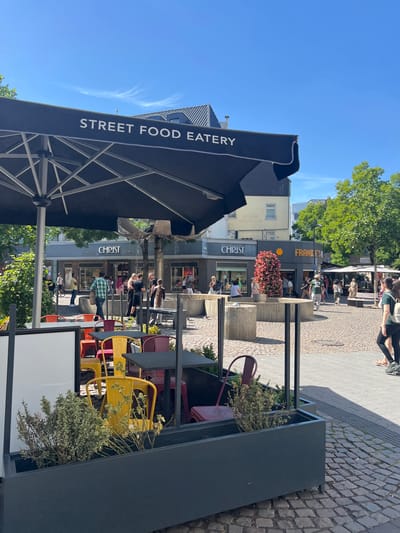 The image captures a sunlit street scene in Aachen, Germany, likely during midday given the bright sunlight and clear blue sky with a few wispy clouds. The perspective is from a slightly elevated viewpoint, looking down onto an outdoor seating area and the street beyond.
In the foreground, a large black umbrella with the white text "STREET FOOD EATERY" emblazoned on its side dominates the left side of the frame. Adjacent to it is a dark grey planter filled with greenery. Behind this, a row of shops lines the street. One shop clearly visible has a sign reading "FRANKFURTER" with a stylized orange logo. Another shop's sign displays "CHRIST SCHMUCK UHREN," indicating it sells jewelry and watches.
The street is paved with cobblestones. Several people are visible, both sitting at outdoor tables and walking along the street. Some are patrons of the eateries, while others appear to be shoppers or passersby. The people are casually dressed, suggesting a relaxed, everyday atmosphere. The overall mood is bright and cheerful, amplified by the sunny weather. Several colorful metal chairs are scattered around the outdoor seating areas, adding a touch of vibrancy to the scene. The presence of trees with lush green foliage suggests a warm season, likely late spring or summer.
