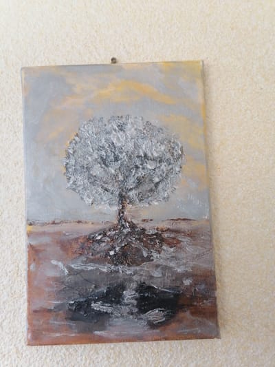 A textured painting depicts a stark, white, bare tree with prominent roots against a muted background of grey and yellow sky and brown earth. The tree's roots spread out across the lower portion of the canvas, extending into a dark, reflective surface that suggests water or wet ground. The sky above is rendered with broad strokes of grey and touches of pale yellow, creating a somber atmosphere. The painting is hanging on a textured wall, as evidenced by the raised surface and a small nail visible at the top edge of the artwork. There are no people or animals present in the image. The location in the city of Sheptytskyi, Ukraine is not discernible from the provided image.