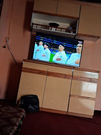 The image shows a television screen displaying a soccer match, likely the FIFA World Cup given the "FUTBOL" graphic. The players on screen are wearing white jerseys. The television is mounted above a wooden cabinet with several drawers and doors. To the left of the cabinet, a black bag rests on the floor next to a patterned rug. A wall socket with a plug is visible on the wall to the left of the television. The overall setting appears to be a living room or common area. The image was taken in Kahramon, Uzbekistan.