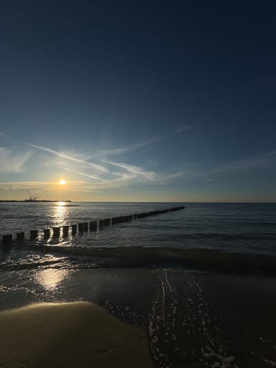 The image depicts a sunset at the beach in Ustka, Poland. The sun, a bright golden orb, is positioned low in the sky, casting a shimmering reflection on the calm sea. A line of weathered wooden posts, partially submerged in the water, extends into the distance. In the background, a crane and other industrial structures are visible along the coastline, suggesting a harbor or port area. The sky is a gradient of deep blue, transitioning to lighter shades near the horizon, with wispy white clouds streaked across it. A flock of birds is seen in the upper right portion of the sky. The foreground shows a sandy beach with gentle waves lapping at the shore, leaving foamy trails on the wet sand. The overall scene is one of serene natural beauty combined with industrial elements.