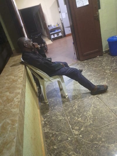 An adult male is seated in a white plastic chair in an indoor environment in the city of Nibo, Nigeria. The man, appearing to be an older Black male with a bald or closely shaven head, wears a dark long-sleeved shirt, dark trousers, and dark slip-on sandals. He is leaning back in the chair with his legs extended forward. His left hand is raised to his mouth or chin area. The floor is tiled with brownish-grey marble-patterned tiles showing reflections. To the immediate left is a light brown tiled wall or partition. In the background, through an open dark brown door, another room or hallway is visible. This room contains a partially visible dark wheelchair, a multi-tiered shelving unit holding various items, and a second partially open dark brown door with a document attached. To the right of the seated man and the doorway, a blue plastic waste bin with a lid is positioned against a wall.