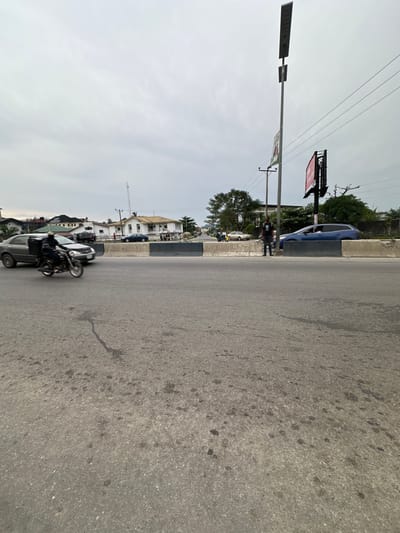 A street scene depicts a light gray asphalt road, slightly cracked, stretching horizontally. A curb separates it from a sidewalk with a few pedestrians, cars, and a motorcycle. A street light is on the left. Further off, a white building with a blue car parked in front is visible. A billboard stands tall with power lines running across the top of the frame. The sky is overcast.