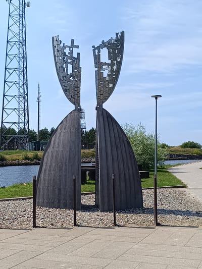 The image shows a public monument in Pāvilosta, Latvia. The monument is composed of two large, curved, dark gray, fluted concrete structures that meet at the top, creating a central gap. Above this, two abstract metal sculptures, resembling shields with cut-out geometric patterns, are positioned to form a mirrored pair, further widening the gap. The monument stands on a bed of light-colored gravel, demarcated by short, dark metal posts. In the foreground, a paved walkway made of light gray concrete slabs extends across the frame. Behind the monument, a body of water, likely a river or sea, is visible, with a grassy embankment and trees on the far side. To the left of the monument, a tall, lattice metal telecommunications tower with an antenna on top is prominent against a clear blue sky with scattered clouds. Another smaller telecommunications tower is visible behind the monument. A single, slender lamppost stands to the right of the monument. The overall setting appears to be a waterfront area, possibly a promenade or park. There are no people visible in the image.
