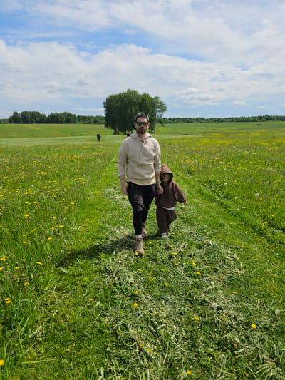 A man and a child are standing in a grassy field with scattered yellow flowers and dandelions under a partly cloudy sky. The man is wearing a light-colored hoodie, dark pants, and sunglasses. The child is wearing a brown hooded outfit. A line of trees is visible in the background. The photo was taken in Uyut, Russia.