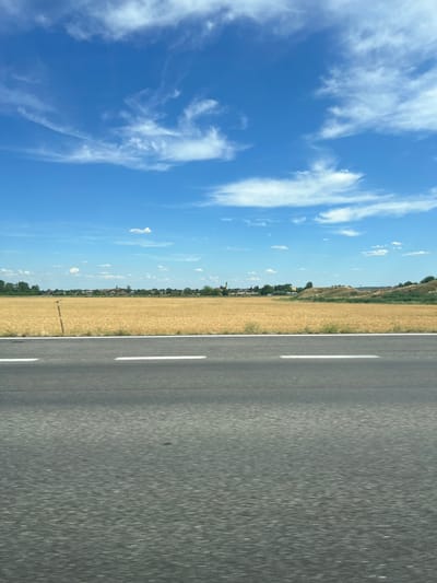 The image shows a view of a rural landscape with a blue sky and scattered white clouds. In the foreground, there is a dark asphalt road with two white lines. Beyond the road, there is a field of golden grain. In the distance, a small town or village is visible with buildings and trees. The scene suggests a sunny day in a countryside setting, potentially in the vicinity of Veľký Biel, Slovakia, given the context provided. The image appears to be taken from a moving vehicle, as indicated by the motion blur on the road. There are no people or specific actions depicted.