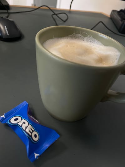 An individual's desk setup features a computer mouse, a mug of coffee with frothed milk, and a single wrapped Oreo cookie. The mouse is black and has the "Tecknet" logo visible. The mug is a light olive green color and is filled with a creamy coffee beverage. The Oreo cookie is in its blue wrapper, with the iconic "OREO" logo prominently displayed. The scene is set on a dark gray desk, and the lighting suggests an indoor environment, likely an office or home workspace. There is no indication of the specific location within Morsbach, Germany.