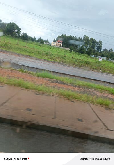 The image captures a static, outdoor scene in Sigona ward, Kenya, viewed through a transparent surface bearing multiple water droplets. The foreground presents a light brown paved surface, possibly a sidewalk or shoulder, exhibiting dark linear marks and an adjacent dark, reflective area indicative of standing water. Beyond this, a two-lane asphalt road, dark and wet with visible surface irregularities, extends horizontally across the midground. Flanking the road are sections of reddish-brown soil interspersed with sparse green grass. The background reveals a gently sloping green hillside covered in grass and numerous trees. Several horizontal, thin dark lines, identified as utility wires, traverse the upper portion of the frame. On the hillside, multiple wooden fence posts are arranged in a line. Further up the slope, a two-story building with light-colored walls and a reddish-brown tiled roof is partially visible amidst a cluster of trees, displaying decorative architectural elements. A white rectangular sign with red markings is situated on the far right side of the hillside. The sky is uniformly overcast and light grey. No human figures are present.