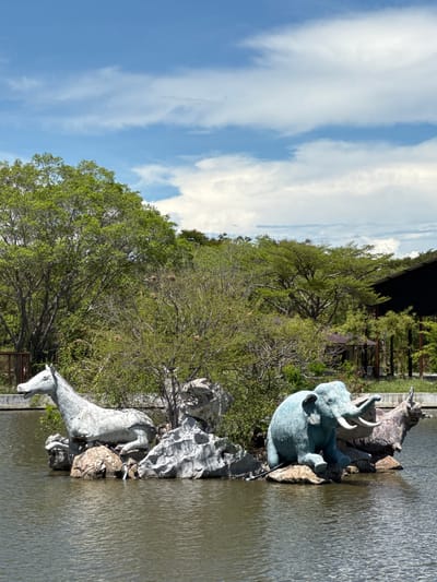 This outdoor shot in Bang Pu Subdistrict Municipality, Thailand, features animal sculptures within a body of water. The sculpture on the left appears to depict a horse or pony, while an elephant sculpture sits towards the upper right of the frame. Both figures are set amongst rocks and vegetation. The water surface shows subtle ripples. Behind the sculptures are trees and foliage, with a partial view of what appears to be a building or structure. Above, a blue sky is visible with scattered white clouds.