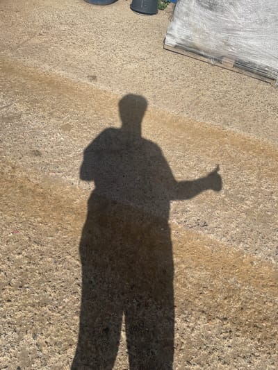 The image shows the shadow of a person on a concrete surface. The shadow is cast from the left, indicating the sun is to the left of the frame. The person's shadow is giving a thumbs-up gesture. The concrete surface appears to be outdoors, with some patches of wetness. In the background, to the right, is a pallet wrapped in clear plastic, and to the left, partially visible, are some dark cylindrical objects, possibly containers or barrels, and some green foliage. The overall scene suggests a sunny day in an outdoor area, potentially a storage or work yard. The location specified, Терволово, Russia, provides a geographical context for the scene.