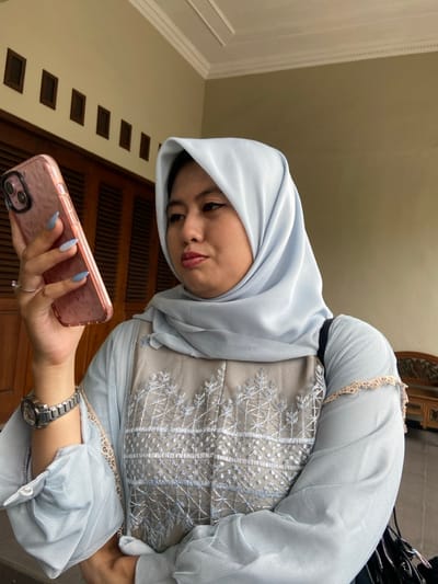 In Ketegan, Indonesia, an adult female is captured in a close-up, chest-level view within an indoor or covered outdoor residential setting. She is wearing a light blue hijab and a matching long-sleeved garment featuring a central beige panel with intricate white embroidered patterns. Her left wrist displays a silver-toned metal watch with a round face. In her left hand, which has light blue gradient nail polish, she holds a rose-gold colored smartphone with a textured case, angled downward. Her head is tilted slightly, and her gaze is directed towards the phone's screen. Her facial features include dark eyeliner and red lipstick. A black strap, consistent with a bag, is visible over her right shoulder. The background includes a light beige wall, a dark brown slatted wooden door, small rectangular wall decorations, and white ceiling molding. A piece of carved wooden furniture is partially visible to the far right.