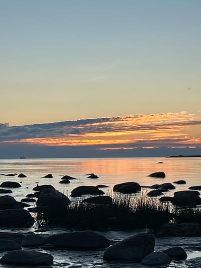 The image depicts a serene coastal scene at dusk in Haabneeme alevik, Estonia. The sky is a gradient of pale blue and soft orange, with wispy clouds catching the last rays of the setting sun. The sea is calm, reflecting the colors of the sky. Numerous dark, smooth rocks are scattered along the shoreline and in the shallow water, some partially submerged. In the distance, a faint silhouette of a ship can be seen on the horizon. The overall impression is one of tranquility and natural beauty.