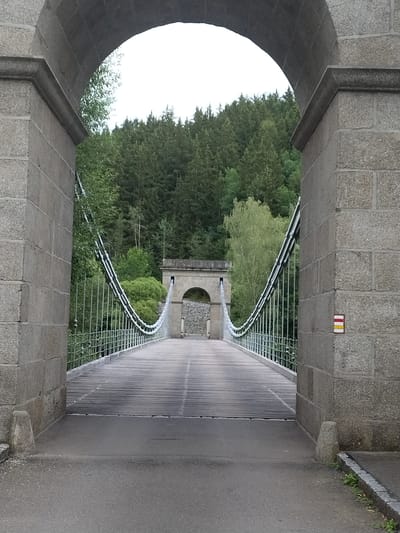 The image shows a view through a stone archway framing a suspension bridge. The bridge has wooden planks for its surface and is suspended by steel cables attached to stone pillars on either side. In the distance, another arched stone structure can be seen at the end of the bridge. The background is filled with a dense forest of evergreen trees. On the right-hand stone pillar, there is a small trail marker with red, yellow, and white stripes. The scene is set in Slavňovice, Czechia, as indicated by the presence of the trail marker, which is a common marking for hiking trails in the Czech Republic. The lighting suggests it is daytime, possibly overcast. There are no people visible in the image.